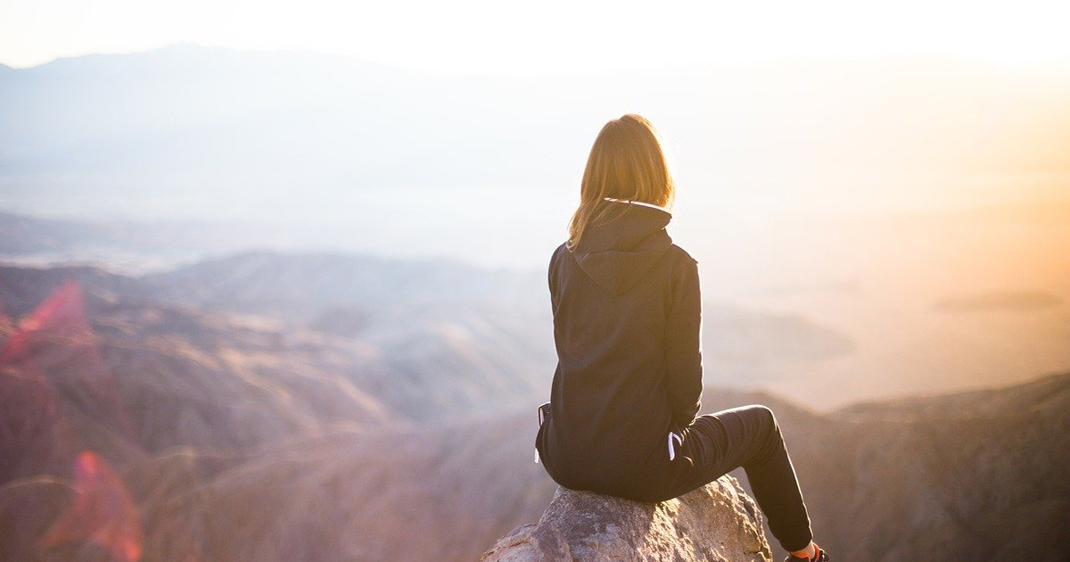 Looking out across a vast mountain vista, a woman sits high on a rock.