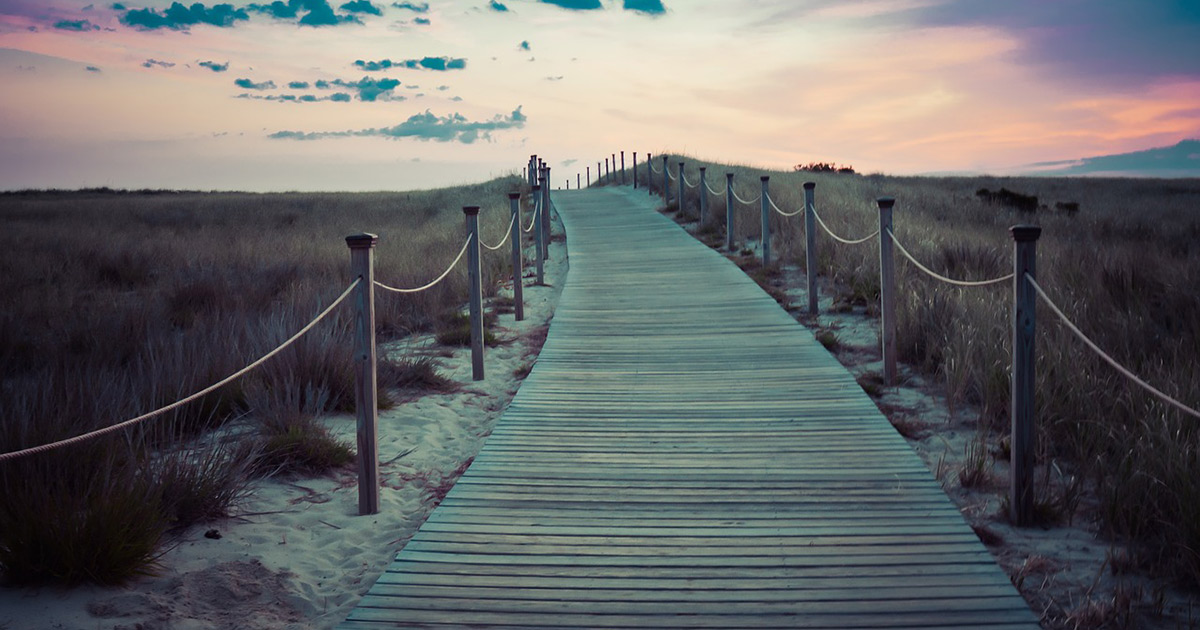 The sun sets in the distance as a wooden path leads through a sandy bluff towards it.