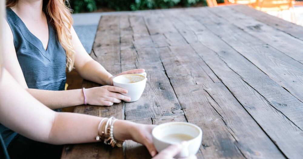 Two women talk at a wooden table, with coffee.