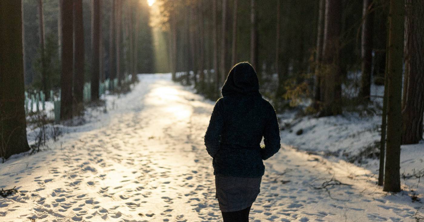 A woman wearing a gray hoodie walks alone along a snowy path in the woods.